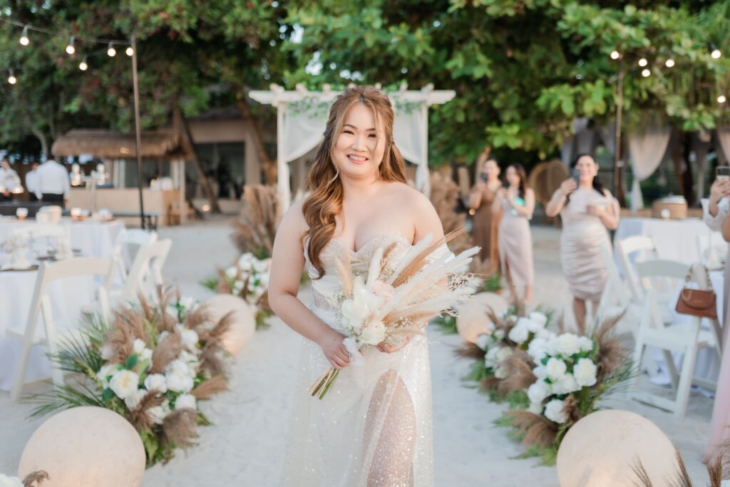 Bride smiling while holding a dried-floral bouquet, walking a sandy aisle lined with pampas grass and white blooms at a beach reception.