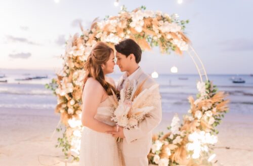 Newlywed couple touching foreheads beneath a circular pampas-and-rose arch at twilight on the beach, ocean and boats in the background.
