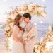 Newlywed couple touching foreheads beneath a circular pampas-and-rose arch at twilight on the beach, ocean and boats in the background.