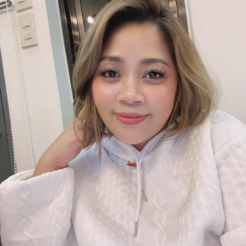 A woman with short, light brown hair smiles softly at the camera while wearing a textured white hoodie, seated indoors.