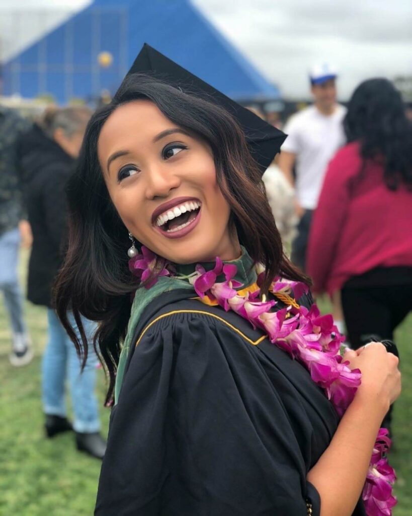 Dr. Kathleen Besas smiling joyfully at her graduation wearing a black cap and gown with a purple orchid lei.