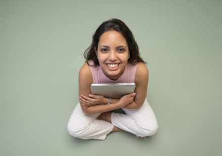 Nancy Gupta smiling and holding a tablet while sitting cross-legged against a sage green background