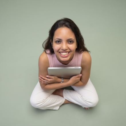 Nancy Gupta smiling and holding a tablet while sitting cross-legged against a sage green background