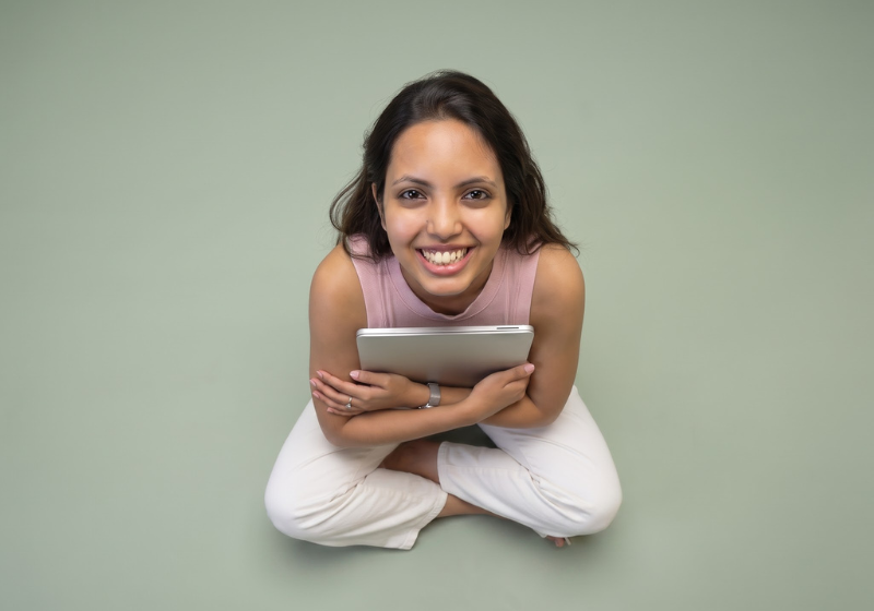 Nancy Gupta smiling and holding a tablet while sitting cross-legged against a sage green background