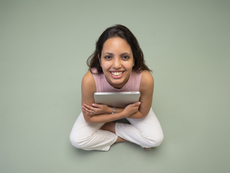 Nancy Gupta smiling and holding a tablet while sitting cross-legged against a sage green background